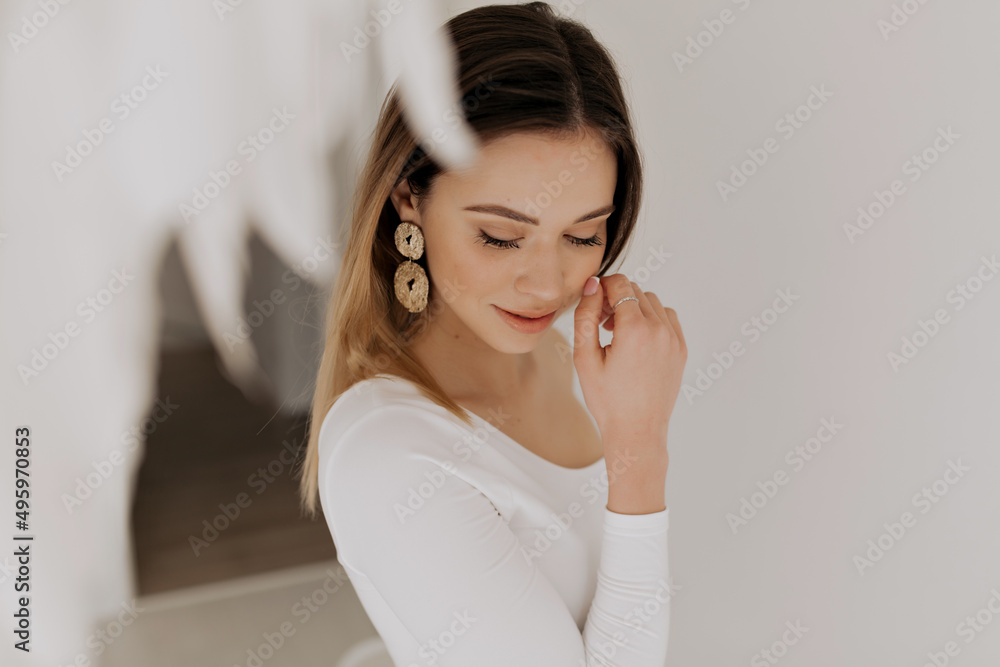 Pretty tenseness woman with nude make up wearing golden earrings is touching her cheek and smiling with closed eyes over isolated background Pretty tenseness woman with nude make up wearing golden earrings is touching her cheek and smiling with closed eyes over isolated background