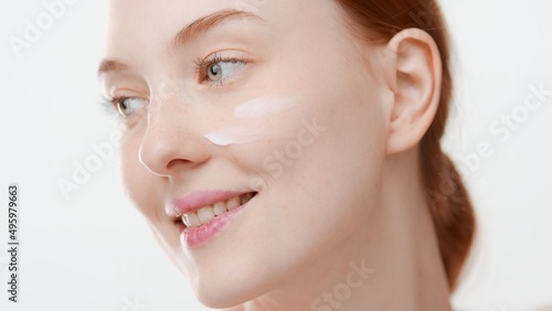 Pretty ginger young Caucasian woman with cream smears on her cheek smiles and looks aside on light grey background   Face cream commercial concept