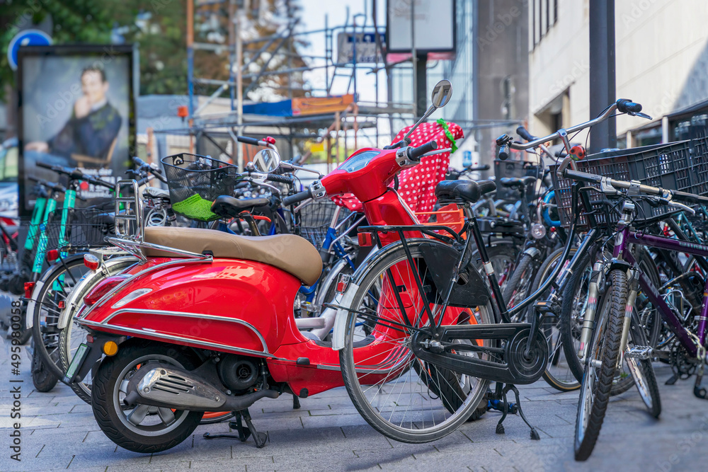 Vivid red scooter standing near many different bicycles on parking