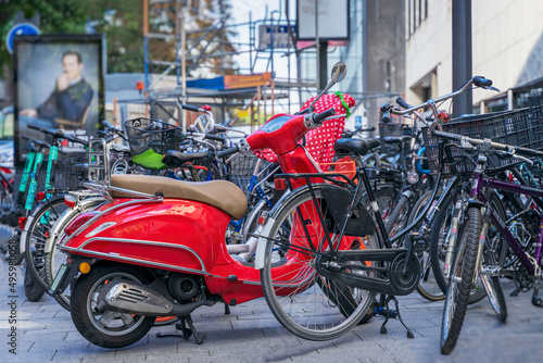 Vivid red scooter standing near many different bicycles on parking