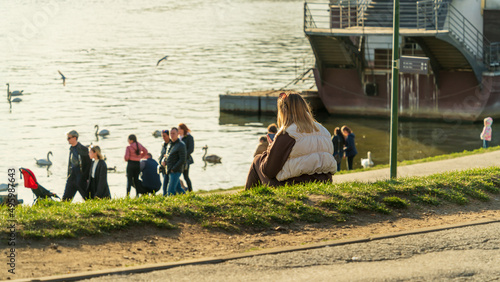A woman with a phone in her hand sits on the green shore of the embankment, looking at waterfowl in the rays of the evening sun. Back view without a face
