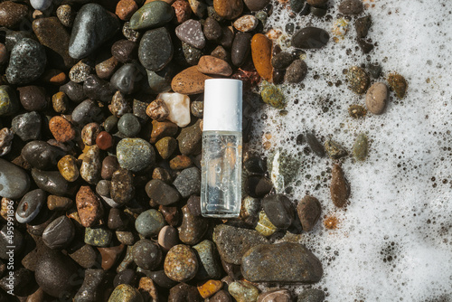 Beauty product transparent bottle on wet beach stones and surrounded by sea foam from the right. Fresh ocean scented product mock up. 