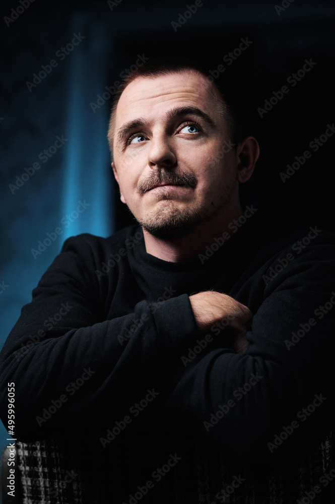 Portrait of a pensive guy in a black jacket, in a dark room on a chair. He crossed his arms in front of him and looked away. Blue eyes Portrait of a pensive guy in a black jacket, in a dark room on a chair. He crossed his arms in front of him and looked away. Blue eyes