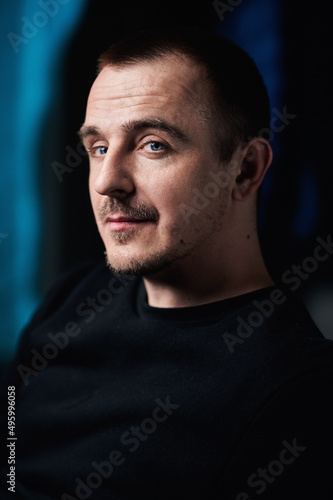 Portrait of a guy in a black jacket, in a dark room. Sly looks away. Blue eyes