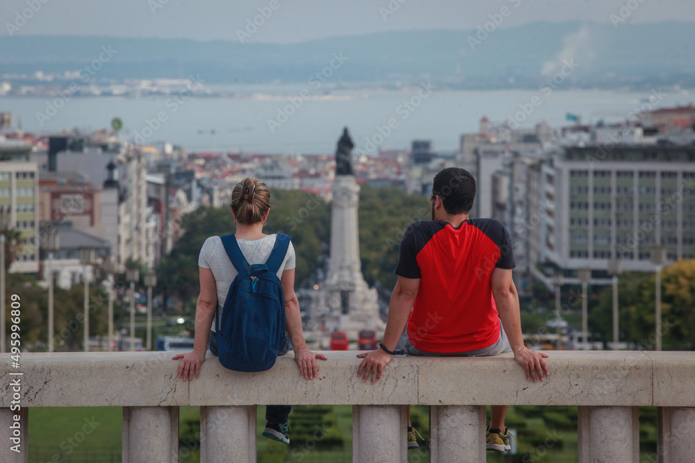 Happy tourist couple sitting at Miradouro Park Eduardo VII in Lisbon, Portugal, Europe, looking towards gardens in the park with Marquess of Pombal Statue and Avenida de Liberdade in a background Happy tourist couple sitting at Miradouro Park Eduardo VII in Lisbon, Portugal, Europe, looking towards gardens in the park with Marquess of Pombal Statue and Avenida de Liberdade in a background