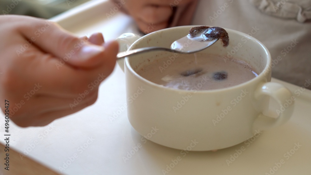 Closeup plate flakes with milk mom feed young baby in white feeding up high chair, kid is trying to eat himself, child with food stained face, little Girl eating porridge with spoon. dairy products Closeup plate flakes with milk mom feed young baby in white feeding up high chair, kid is trying to eat himself, child with food stained face, little Girl eating porridge with spoon. dairy products
