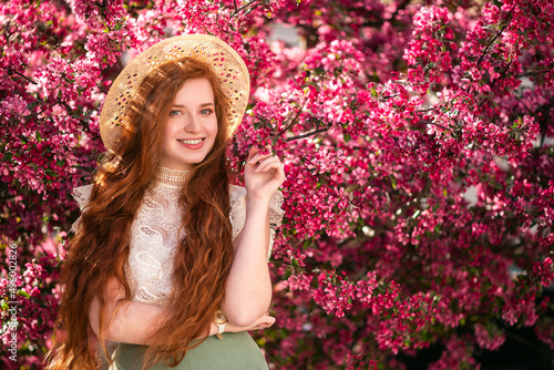 Beautiful fashionable happy smiling redhead, freckled woman with long natural curly hair, wearing straw hat, lace blouse, posing in blooming garden, near blossom trees. Copy, empty space for text