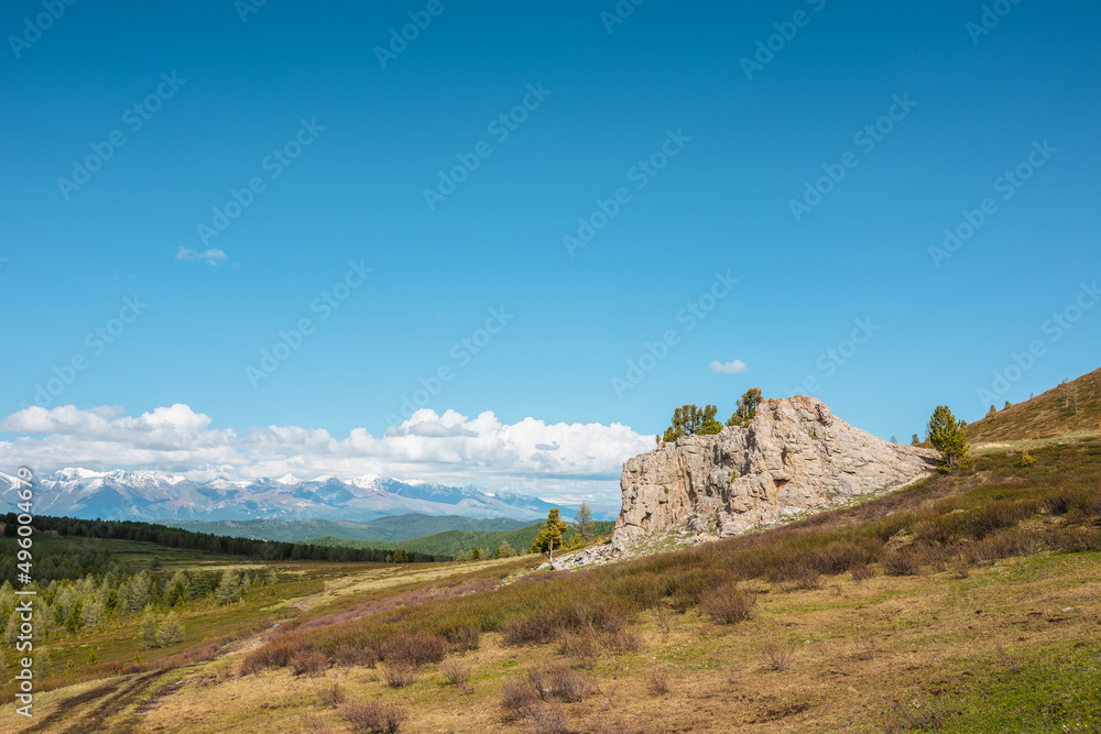 Atmospheric landscape with big rock on hillside with view to sunlit mountain vastness and high snowy mountain range under low clouds line on horizon. Impressive scenery with vast mountains in sunlight