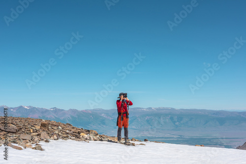 Tourist in red walks on snow mountain near abyss edge on high altitude under blue sky in sunny day. Man with camera on snowy mountain near precipice edge with view to large mountain range in away. © Daniil