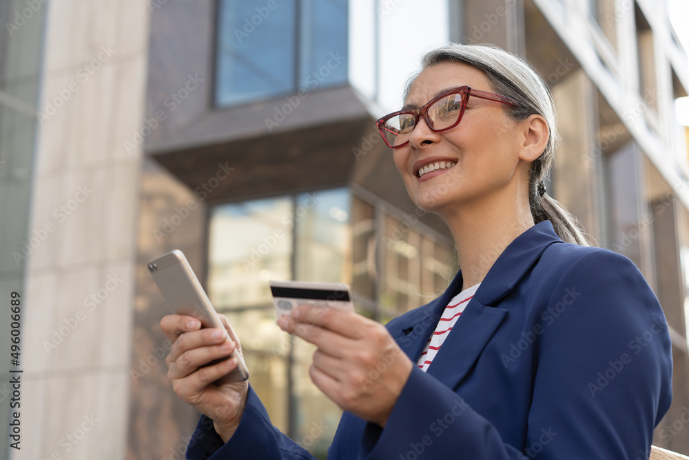 Confident smiling mature businesswoman holding credit card using mobile phone shopping online on the street. Modern technology, e-commerce concept Confident smiling mature businesswoman holding credit card using mobile phone shopping online on the street. Modern technology, e-commerce concept