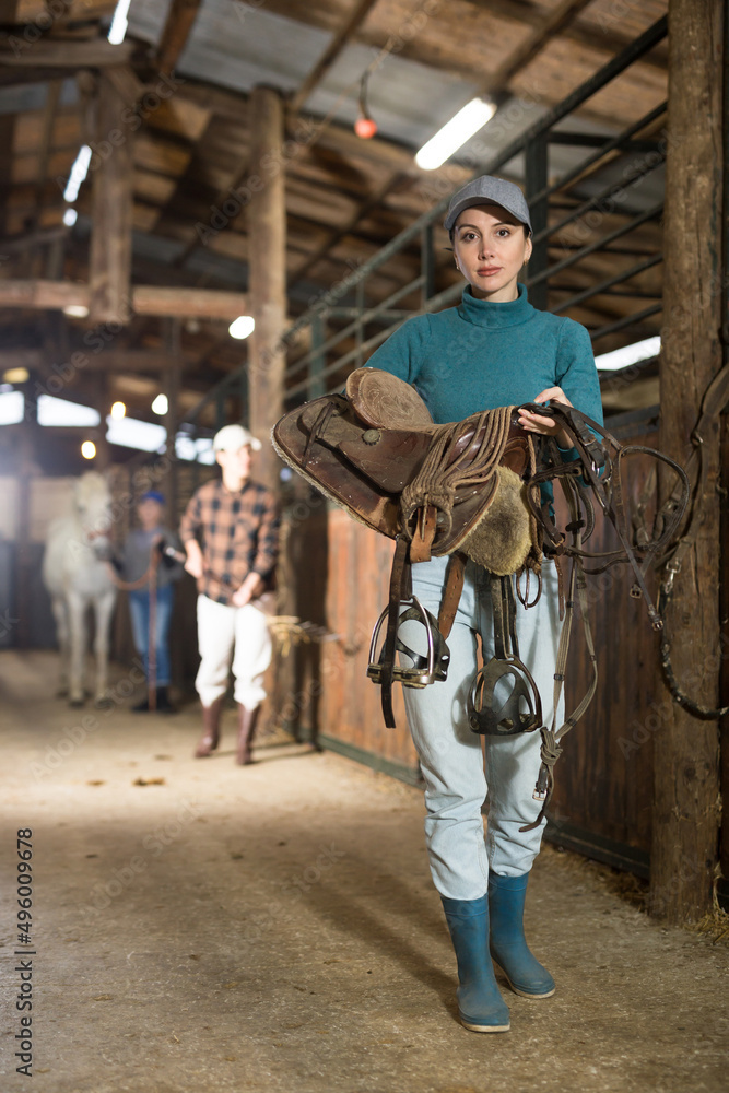 Woman jockey with saddle and bridle in her hands in the stable