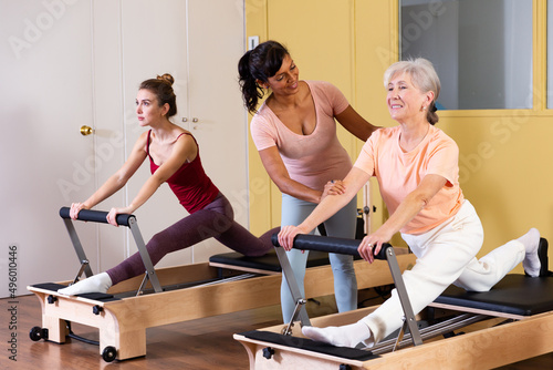 Aged woman doing pilates on reformer in fitness studio with Hispanic female personal trainer controlling movements