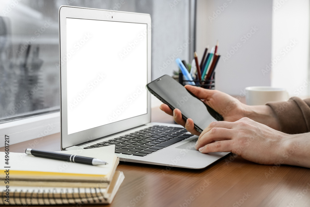 Fototapeta premium Woman checking mobile phone and working with laptop on table near window