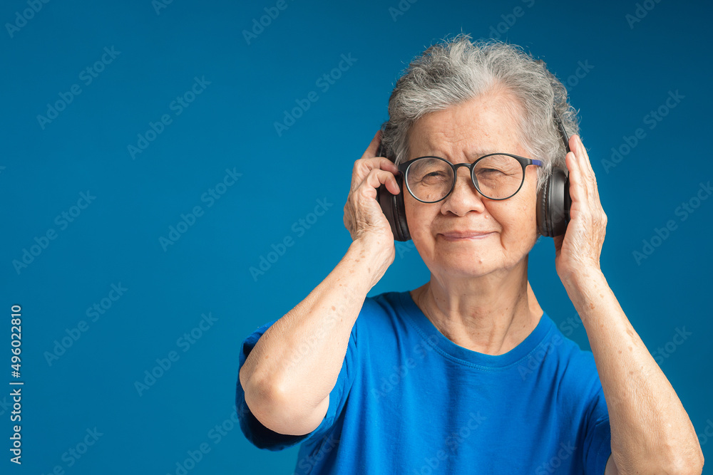 Cheerful senior woman wearing wireless headphones with a smile while standing on a blue background Cheerful senior woman wearing wireless headphones with a smile while standing on a blue background