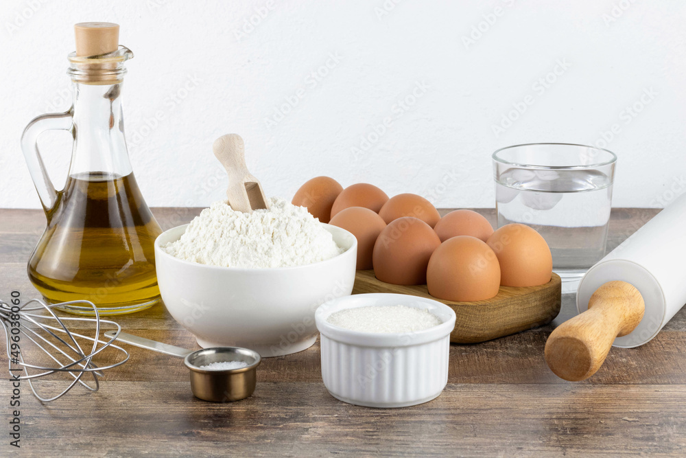 Baking ingredients: butter, flour, sugar, cold water, eggs, baking powder, whisk, rolling pin. Ingredients for making a cake or cupcake on a wooden background, side view. Baking ingredients: butter, flour, sugar, cold water, eggs, baking powder, whisk, rolling pin. Ingredients for making a cake or cupcake on a wooden background, side view.