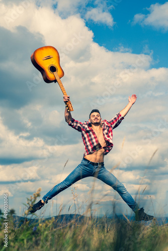 Young excited man jumping with guitar on nature background. Music outdoors. Sexy man with guitar in checkered shirt, hipster camping and hiking.