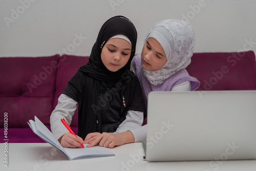 Two child girls in the hijab together learn for school at the home desk. Sister helping little sister to do homework. Young Muslim students have online education.