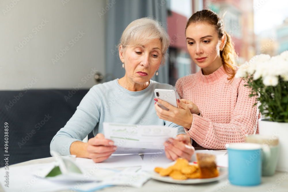 Young woman with smartphone helping grandmother to pay her bills with online payment.