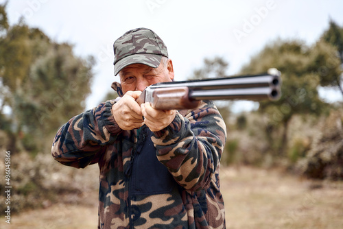 Senior man in camouflage clothes and cap squinting and aiming shotgun while hunting in countryside on autumn day