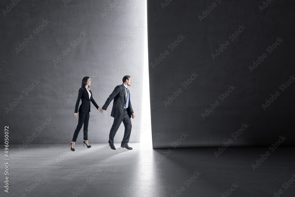 Obraz premium Side view of businessman and woman holding hands and stepping into the light from behind a concrete wall. Success, teamwork and future concept.