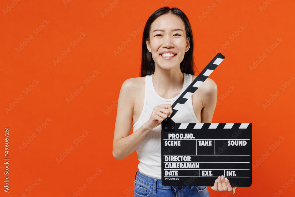 Cheerful trendy fancy charming happy young woman of Asian ethnicity 20s years old in white tank top holding classic black film making clapperboard isolated on plain orange background studio portrait.