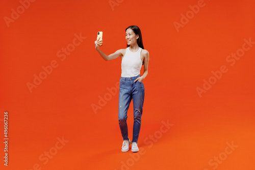 Full size young woman of Asian ethnicity 20s in white tank top get video call use mobile cell phone do selfie talk conducting pleasant conversation isolated on plain orange background studio portrait.