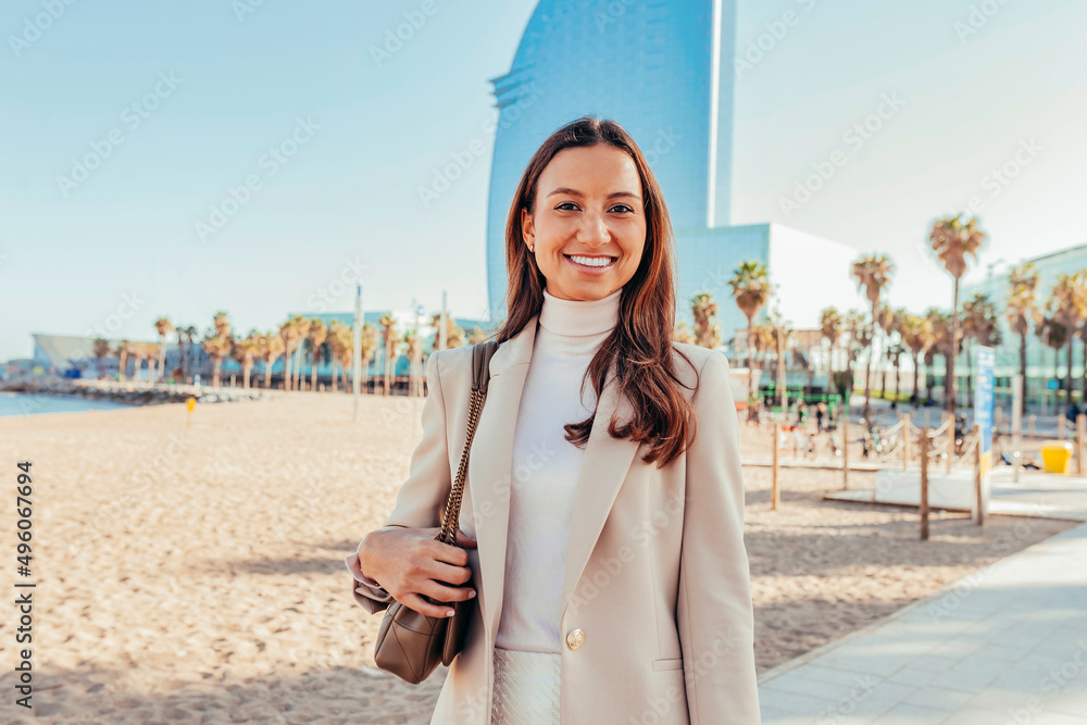 Young happy smiling attractive businesswoman standing staring at camera, at beach of Barcelona city street to office, skyscrapers cityscape and beach with palms background. Copy space. - obrazy, fototapety, plakaty 