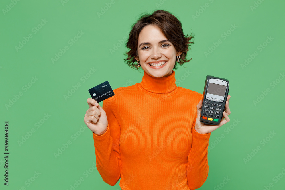 Young smiling woman 20s wear casual orange turtleneck hold wireless modern bank payment terminal to process acquire credit card payments isolated on plain pastel light green color background studio. Young smiling woman 20s wear casual orange turtleneck hold wireless modern bank payment terminal to process acquire credit card payments isolated on plain pastel light green color background studio.