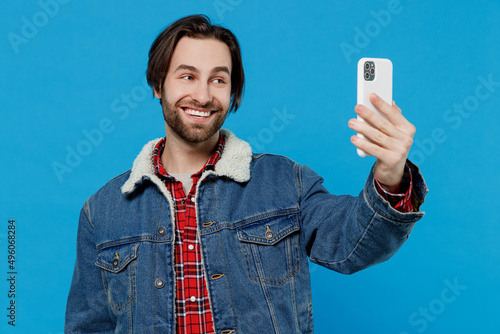 Friendly smiling young brunet bearded man 20s wears warm denim jacket doing selfie shot on mobile cell phone isolated on plain pastel light blue background studio portrait. Winter cold season concept. © ViDi Studio
