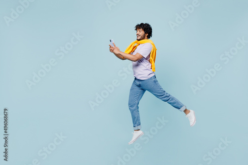 Full size excited young bearded Indian man 20s wears white t-shirt hold in hand use mobile cell phone typing browsing chatting jumping isolated on plain pastel light blue background studio portrait. photo