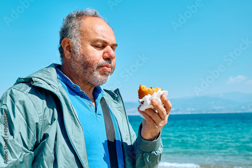 Bearded mature man at spring seaside eating hot palatable arancina (deep fried rice balls with meat). Typical Sicilian street food.