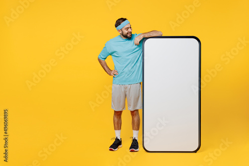 Full length young fitness trainer instructor sporty man sportsman in headband blue tshirt big screen mobile cell phone with copy space mockup isolated on plain yellow background Workout sport concept