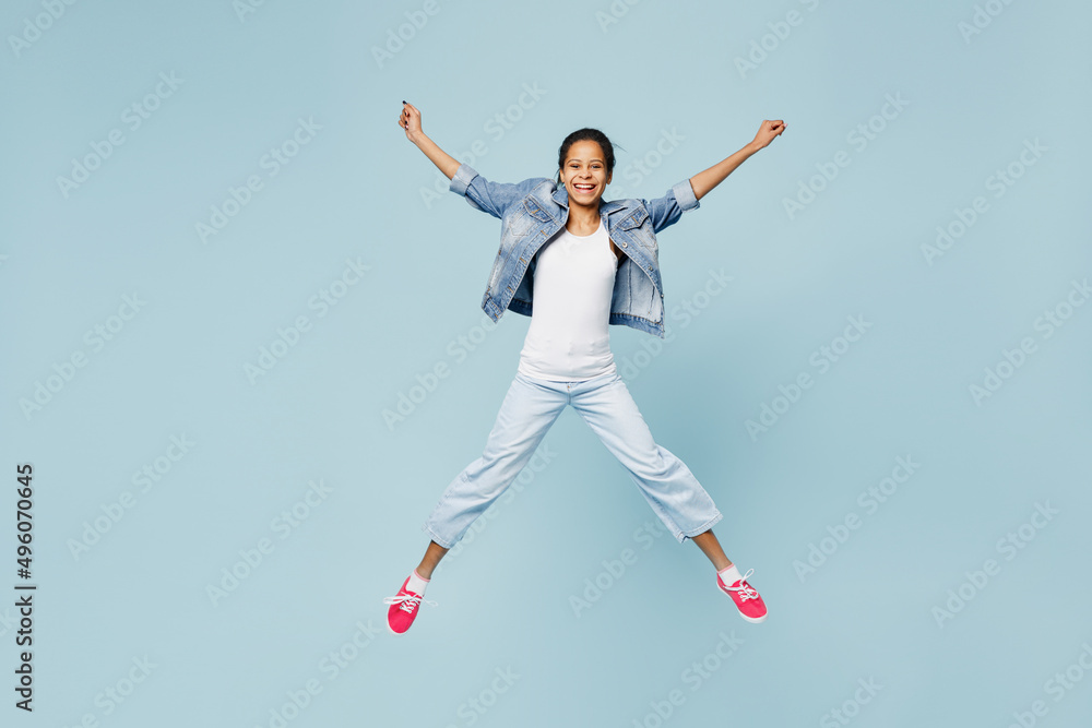 Full size happy little kid teen girl of African American ethnicity 12-13 year old in denim jacket jump high with outstretched hands isolated on pastel plain light blue background. Childhood concept
