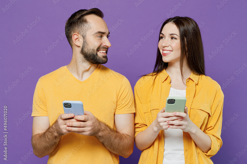 Young smiling happy fun couple two friends family man woman together in yellow casual clothes hold in hand use mobile cell phone chatting look to each other isolated on plain violet background studio. Young smiling happy fun couple two friends family man woman together in yellow casual clothes hold in hand use mobile cell phone chatting look to each other isolated on plain violet background studio.