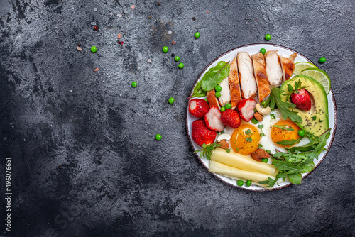 Ketogenic diet. Fried egg, avocado, strawberry, grilled chicken fillet, cheese, nuts and arugula on a dark background, Low carb high fat breakfast, top view
