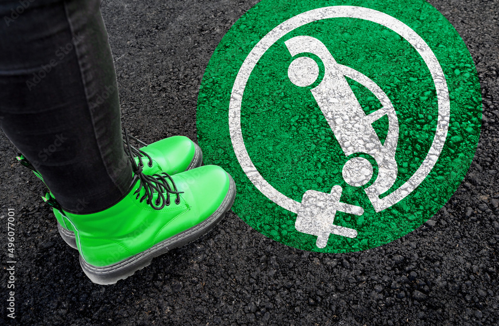 a woman with a boots is standing next to electric car sign on road asphalt  - obrazy, fototapety, plakaty 