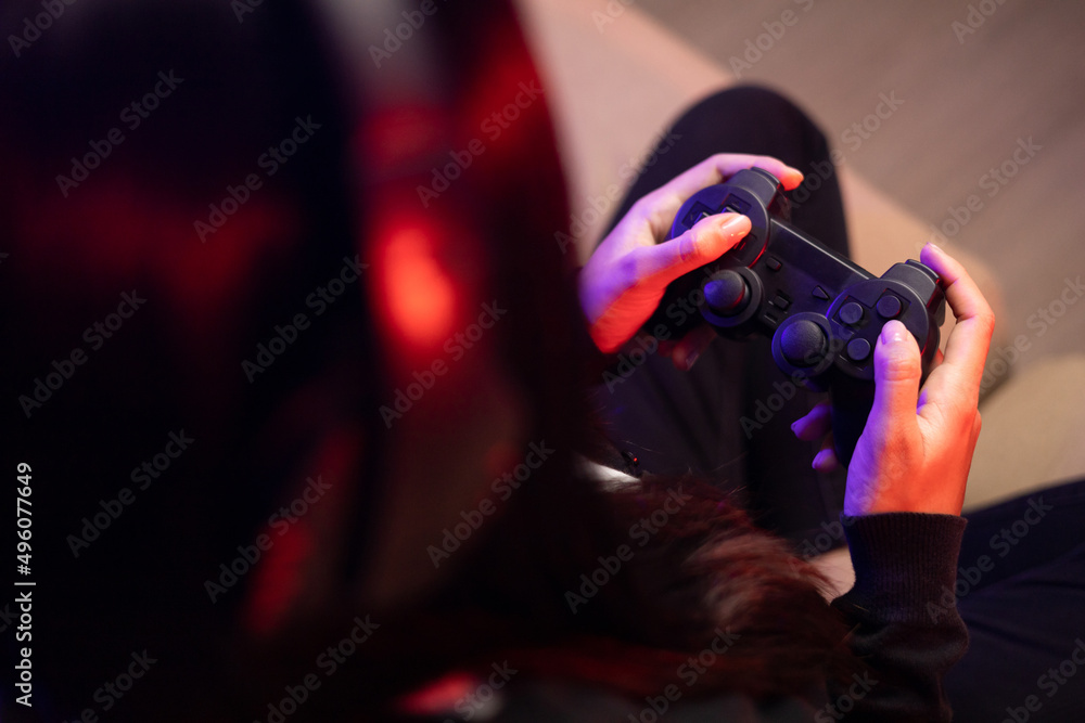 Close up hands Playing video game. Young pretty woman sitting on sofa holding joystick in living room. Female Professional Streamer wearing hoodie playing game online in dark room neon light. Close up hands Playing video game. Young pretty woman sitting on sofa holding joystick in living room. Female Professional Streamer wearing hoodie playing game online in dark room neon light.