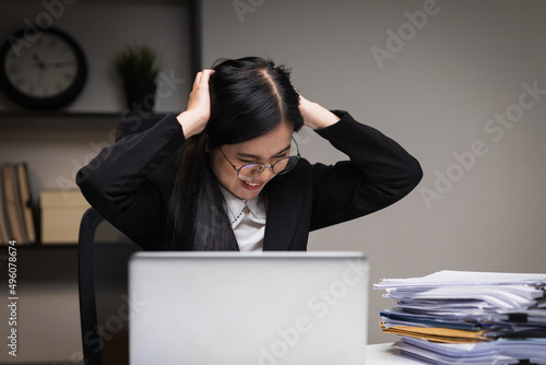Stressed asian business woman working late at night in the office hands on head feeling headache. Tired woman looking at laptop working hard sitting in the dark room office. Overtime concept