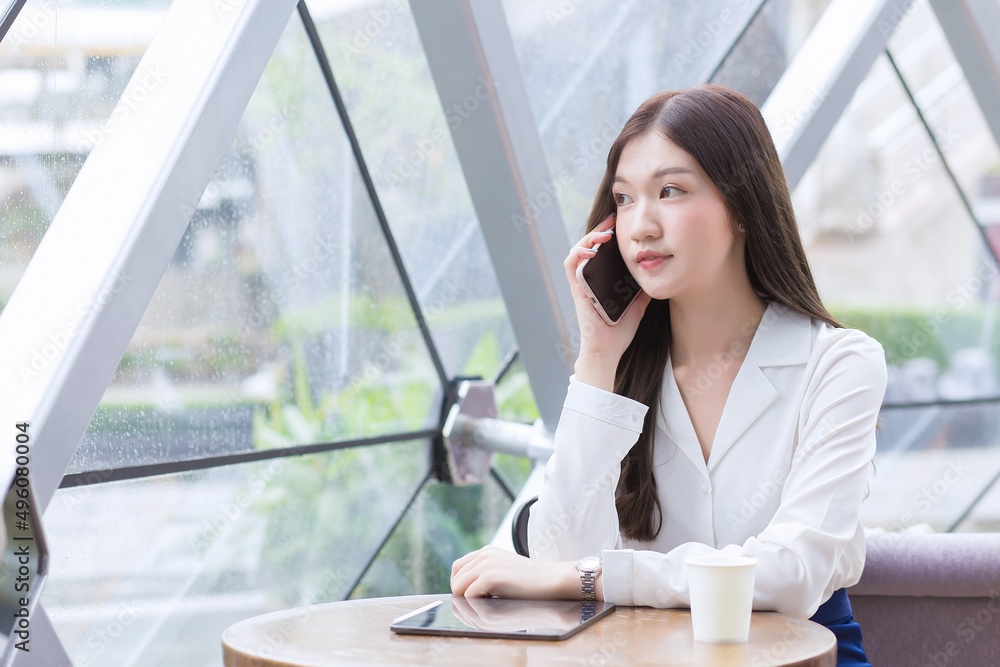Obraz premium Beautiful young Asian professional business female in a white shirt is while using your smartphone in your hand to talk she is sitting happy smile working at coffee shop among business background