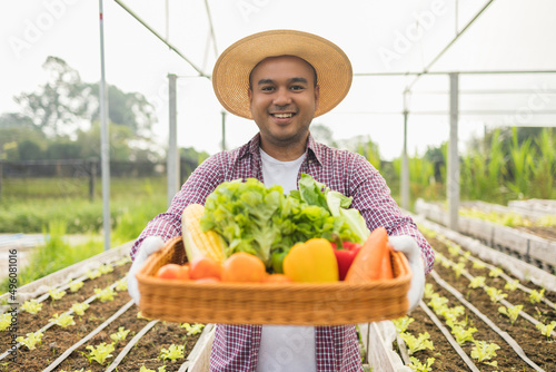 An Asian farmer stands in front of a vegetable garden with a basket full of fresh vegetables. Agriculture until the vegetable harvesting season in the greenhouse