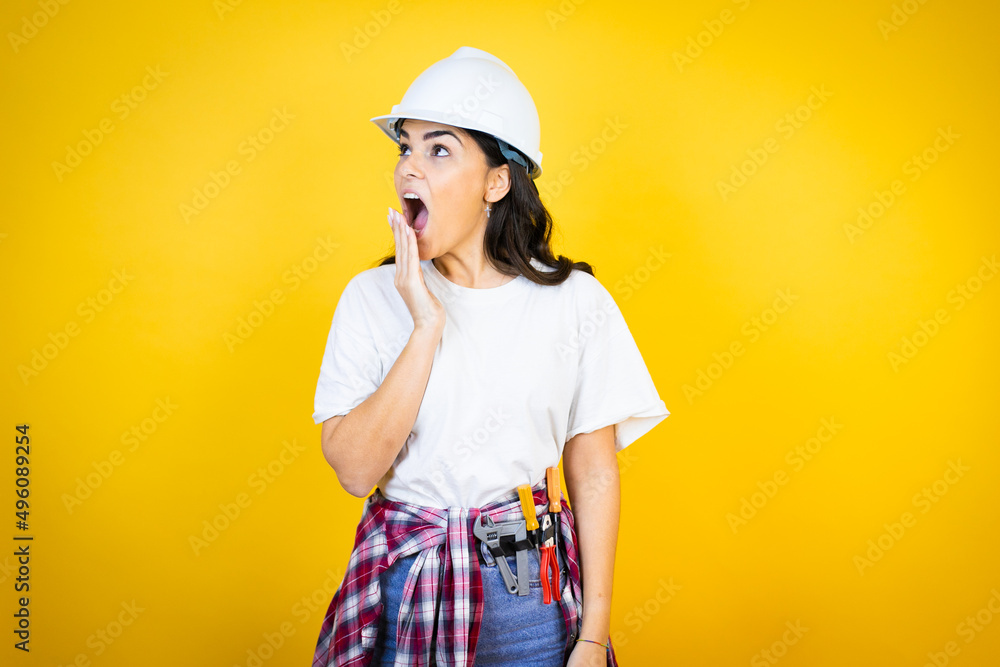 Young caucasian woman wearing hardhat and builder clothes over isolated yellow background covering mouth with hand, shocked and afraid for mistake. surprised expression Young caucasian woman wearing hardhat and builder clothes over isolated yellow background covering mouth with hand, shocked and afraid for mistake. surprised expression