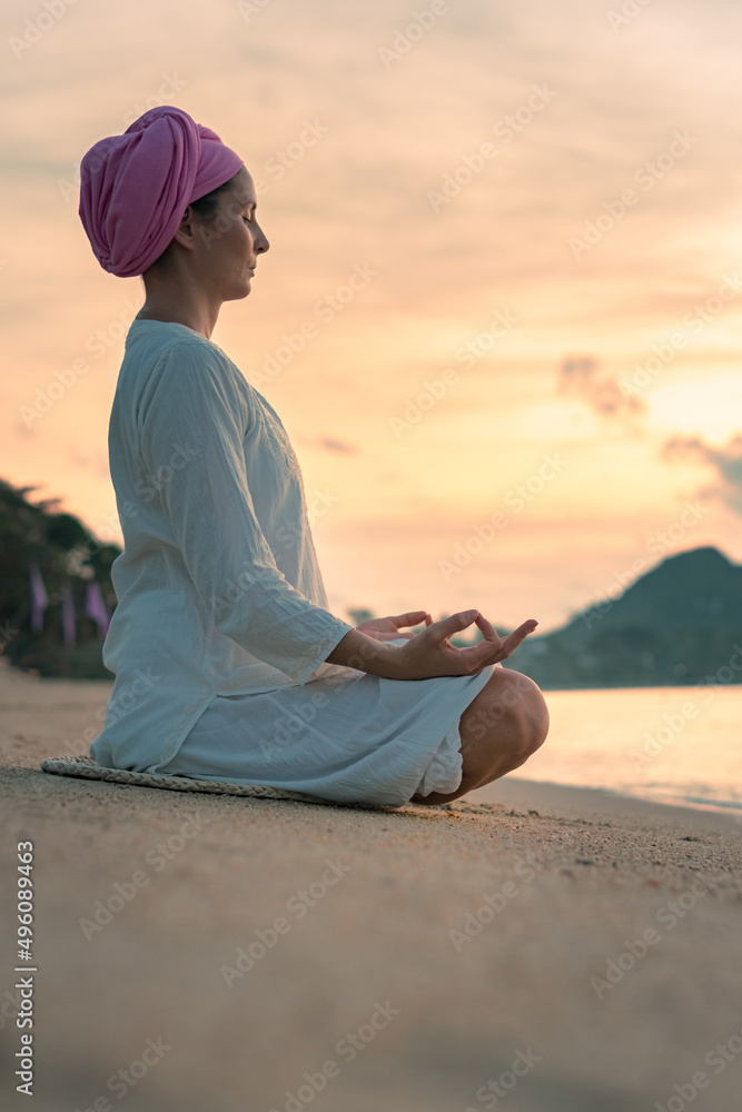 A woman in white clothes and a turban sits by the sea on a sandy beach at dawn on a summer morning and doing yoga exercises . Colorful sky on the background.
