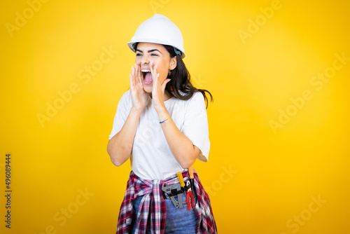 Young caucasian woman wearing hardhat and builder clothes over isolated yellow background shouting and screaming loud to side with hands on mouth