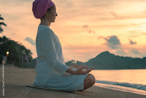 A woman in white clothes and a turban sits by the sea on a sandy beach at dawn on a summer morning and doing yoga exercises . Colorful sky on the background.
