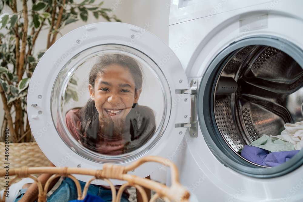 A cute smiling little girl with brown hair and dark eyes looks through the round glass door of the washing machine, watches her mother sorting laundry in the bathroom A cute smiling little girl with brown hair and dark eyes looks through the round glass door of the washing machine, watches her mother sorting laundry in the bathroom