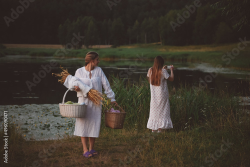 Two beautiful girls in white nightgowns with long beautiful hair on a walk by the lake with a bouquet and baskets in their hands. Warm summer evening. Girls on a picnic. Countryside. photo