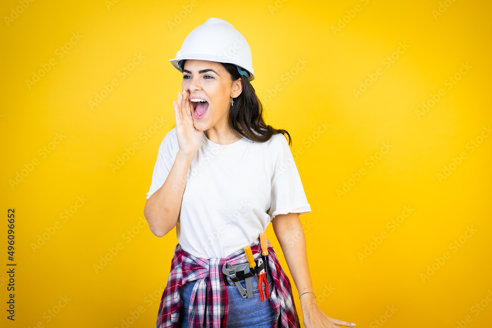 Young caucasian woman wearing hardhat and builder clothes over isolated yellow background shouting and screaming loud to side with hand on mouth Young caucasian woman wearing hardhat and builder clothes over isolated yellow background shouting and screaming loud to side with hand on mouth