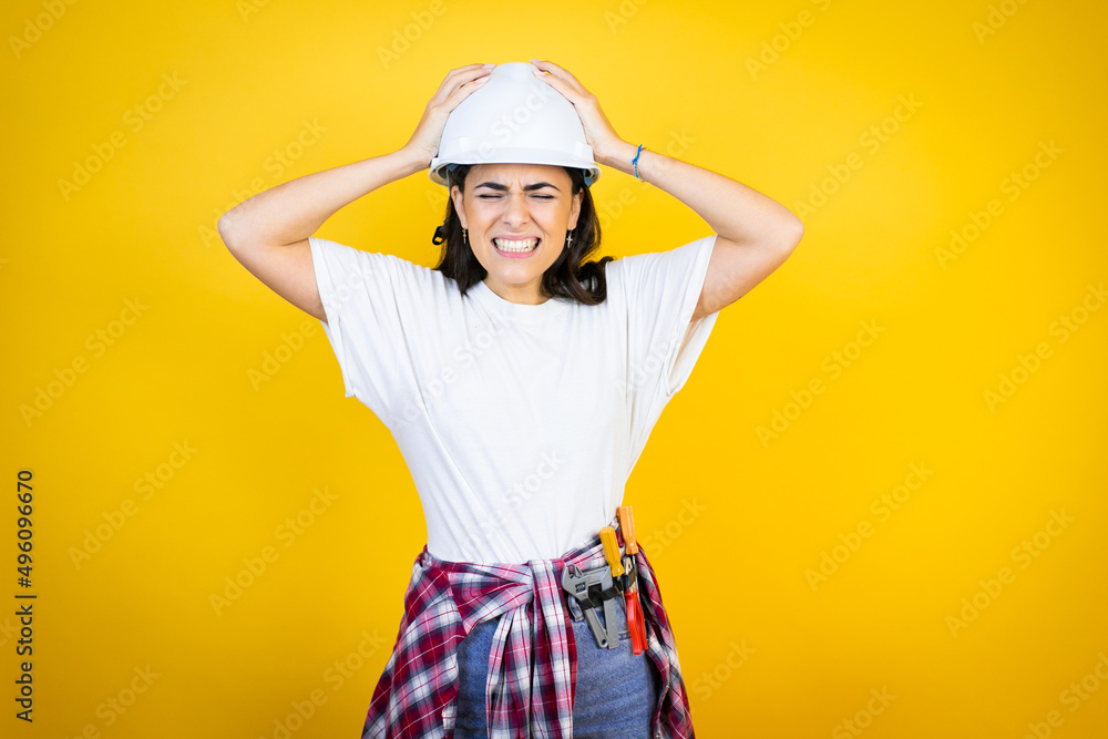 Young caucasian woman wearing hardhat and builder clothes over isolated yellow background suffering from headache desperate and stressed because pain and migraine with her hands on head Young caucasian woman wearing hardhat and builder clothes over isolated yellow background suffering from headache desperate and stressed because pain and migraine with her hands on head