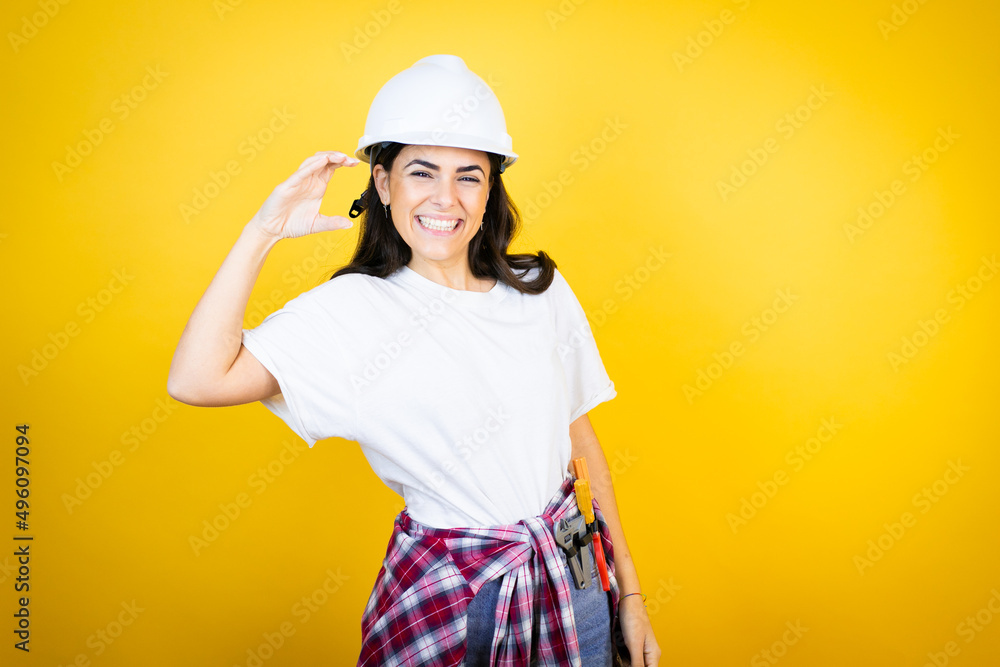 Young caucasian woman wearing hardhat and builder clothes over isolated yellow background smiling and confident gesturing with hand doing small size sign with fingers . Measure concept. Young caucasian woman wearing hardhat and builder clothes over isolated yellow background smiling and confident gesturing with hand doing small size sign with fingers . Measure concept.