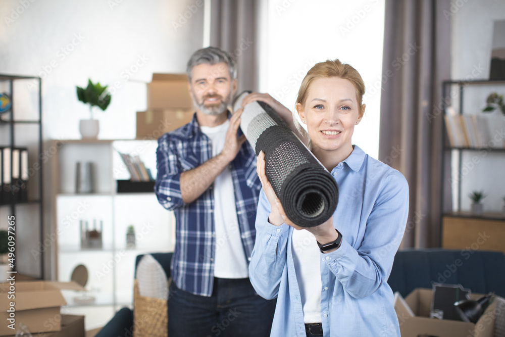 Fototapeta premium Portrait of a smiling middle aged couple carrying rolled rug after moving in a house. Focus on smiling pretty blond woman, holding roll carpet on her shoulder and her pleasant bearded man behind